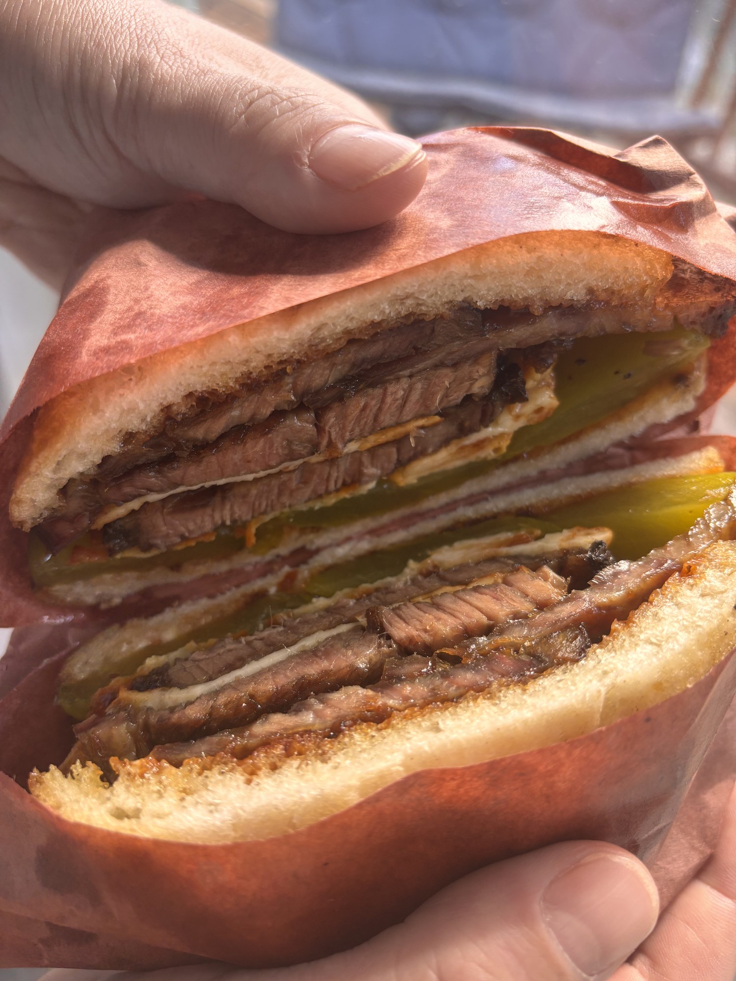 Cut face of the finished sandwich held in one hand: two thick slices of smoked brisket with dark bark edges, a pale layer of fried cotija and bright green pickle spears stacked between two slices of in-house bread, the whole thing cradled in pink butcher paper.