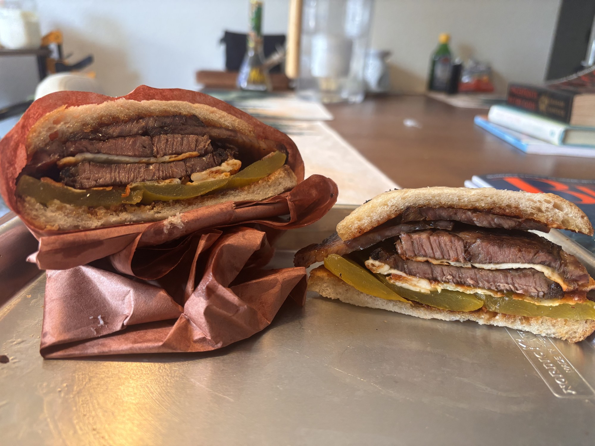 Both halves of the sandwich standing cut-side out on a steel sheet tray, one half still in its butcher paper, showing the brisket and pickle layers from the side.
